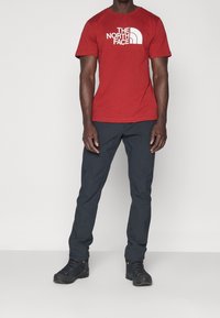 Red cotton t-shirt with white "The North Face" logo, paired with dark gray pants and black hiking shoes. Solid colors, casual fit.