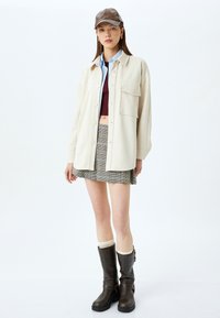 Beige skjorta med knappar över en vinröd crop top, tillsammans med en rutigt minikjol och knähöga bruna stövlar med spännen. En brun keps fulländar looken.