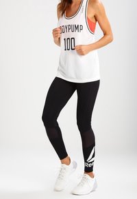 Biały siateczkowy top na ramiączkach z napisem "BODYPUMP 100", nałożony na czerwony stanik sportowy. Czarne legginsy z przezroczystymi akcentami i białym logo Reebok. Białe buty sportowe.