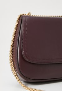 Bordo kožna torbica s zlatnim lančićem, s poklopcem i glatkom текстurom uz ušivene detalje.