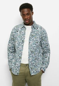 Hemd mit Knöpfen in einem mehrfarbigen Blumenmuster auf hellem Hintergrund, aus weichem Stoff gefertigt. Kombiniert mit einem weißen T-Shirt und grüner Hose.