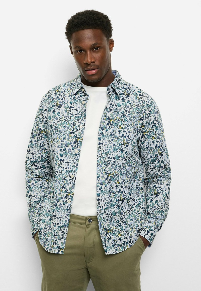 Hemd mit Knöpfen in einem mehrfarbigen Blumenmuster auf hellem Hintergrund, aus weichem Stoff gefertigt. Kombiniert mit einem weißen T-Shirt und grüner Hose.