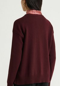 Bordeauxroter Pullover mit lockerer Passform, strukturierter Strickoptik, geripptem Saum und fallenden Schultern. Verfügt über einen gemusterten Kragen darunter.