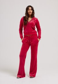 Rode velours zip-up hoodie gecombineerd met bijpassende flared broek. Bevat kangoeroezakken en een trekkoord tailleband. Soepele textuur overal.