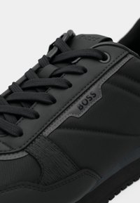 Black BOSS sneaker with textured fabric and leather details, black laces, and brand logo on the side near the laces.