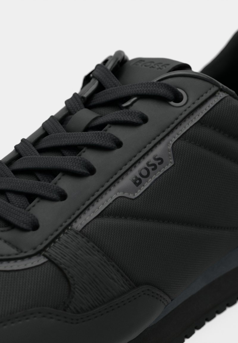 Sneaker BOSS noir avec tissu texturé et détails en cuir, lacets noirs et logo de la marque sur le côté près des lacets.