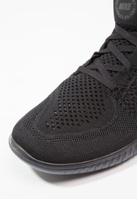 Zapatilla deportiva negra con parte superior de malla texturizada, puntera redondeada y suela de goma con un patrón de desgaste. Marca Nike en el cuello.