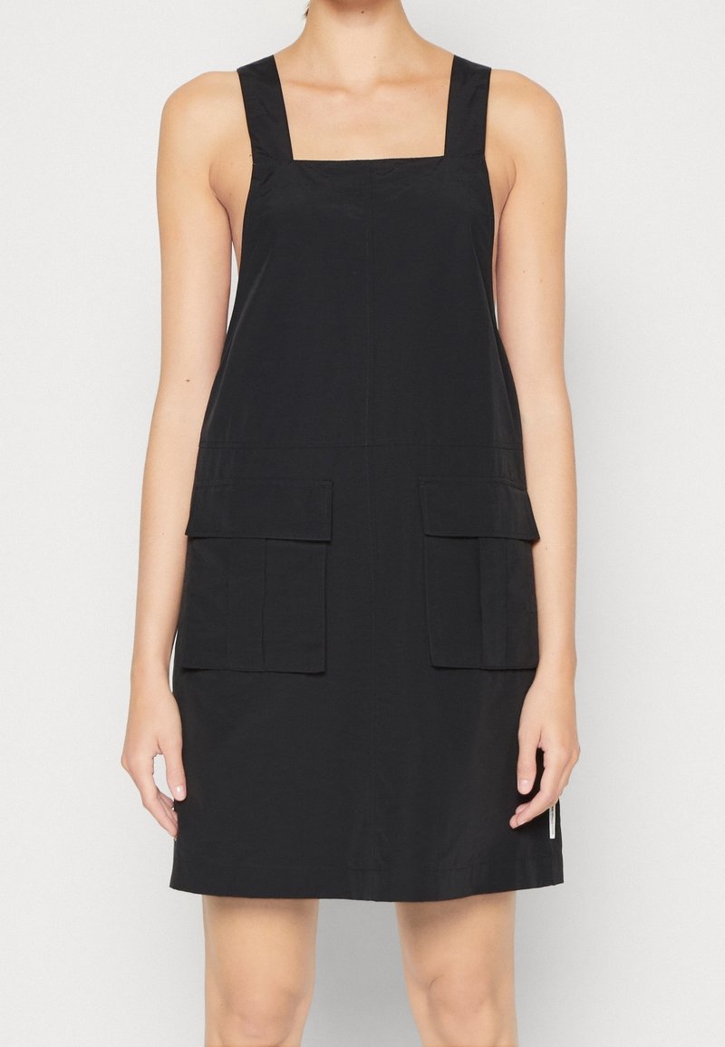 Robe salopette en coton noir avec encolure carrée, larges bretelles, deux poches avant et ourlet droit. Texture lisse avec un design minimaliste.