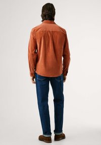 Corduroy overhemd in gedempt oranje, met lange mouwen en knoopmanchetten, gestructureerde rugpaneel, gecombineerd met donkerblauwe spijkerbroeken en bruine schoenen.