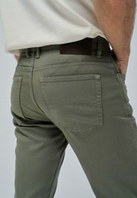 Pantalones de color verde oliva hechos de una mezcla de algodón, con un diseño clásico de cinco bolsillos y una parche de cuero en la parte trasera de la cintura.