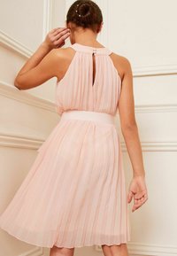 Skládané šaty v jemně růžové barvě, s vysokým výstřihem, klíčovou zadní částí a širokým páskem. Hladká textura s volnou sukní.