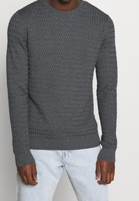 Homme portant un pull en maille texturée gris foncé ajusté et un jean bleu clair, debout devant un fond clair uni.