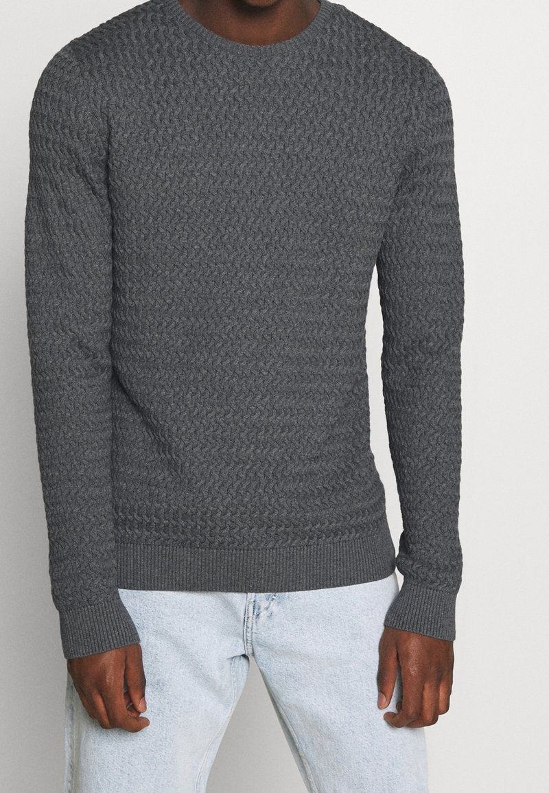 Homme portant un pull en maille texturée gris foncé ajusté et un jean bleu clair, debout devant un fond clair uni.