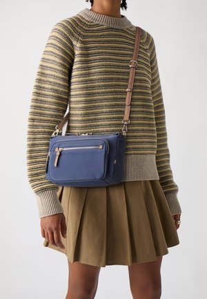 Personne portant un pull en maille rayé et une jupe plissée couleur sable, portant un sac bandoulière bleu marine avec une sangle ajustable tan et une poche avant zippée.