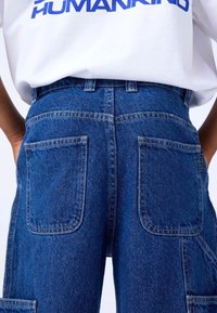 Pantalones cargo de mezclilla en azul oscuro, con una cintura alta, dos bolsillos traseros y un tejido texturizado con detalles de costura visibles.