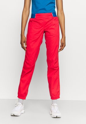 Persoon die felrode broeken met zijzakken, een blauw shirt, grijze sokken en witte sneakers draagt, staande tegen een effen lichte achtergrond.