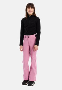 Ružové lyžiarske nohavice majú rovný strih s vysokým pásom a dvoma gombíkmi. V kombinácii s čiernym topom s dlhým rukávom a čiernymi čižmami.