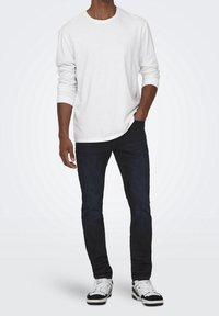 Camiseta blanca de manga larga con cuello redondo, combinada con jeans ajustados oscuros y zapatillas en blanco y negro. Un atuendo simple y casual.