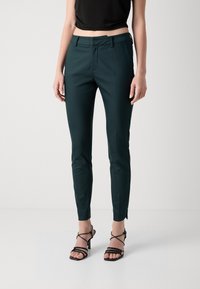 Slim-fit zelené kalhoty s hladkou texturou, se plochým předním dílem, poutky na pásek a zúženými nohavicemi končícími na úrovni kotníků, spárované s páskovými podpatky.