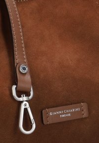 Bolso de ante marrón con una correa de cuero con un cierre de plata y una etiqueta de marca en cuero marrón con letras blancas.