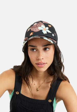 Frau mit langem dunklem Haar, die eine schwarze Blumenmütze, einen schwarzen ärmellosen Overall, ein grünes Oberteil und eine goldene Anhängerkette trägt, leicht seitlich schauend.