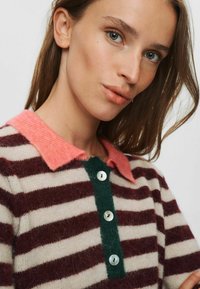 Junge Frau mit blauen Augen und glattem braunem Haar trägt einen gestreiften Strickpullover mit korallenfarbenem Kragen und dunklen Knöpfen.