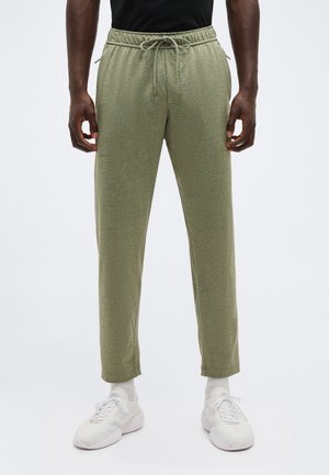 Olívazöld, húzózsinóros jogging nadrágot, fehér zoknit és fehér sportcipőt viselő férfi, egy egyszerű, világos háttér előtt áll.