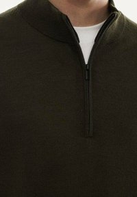 Pull zippé vert olive avec un col montant, fabriqué en tissu tricoté texturé ; doté d'une fermeture éclair frontale et d'une couche intérieure blanche visible au niveau du cou.