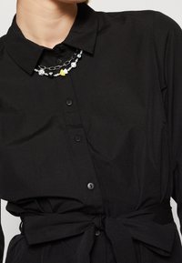 Camicia nera con colletto, dotata di una vita legata e una texture liscia, completata da una collana di perline multicolori.
