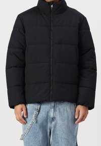 Chaqueta acolchada negra con cuello alto, cierre de cremallera y diseño acolchado. Presenta un ajuste holgado y está hecha de una tela suave e aislante.