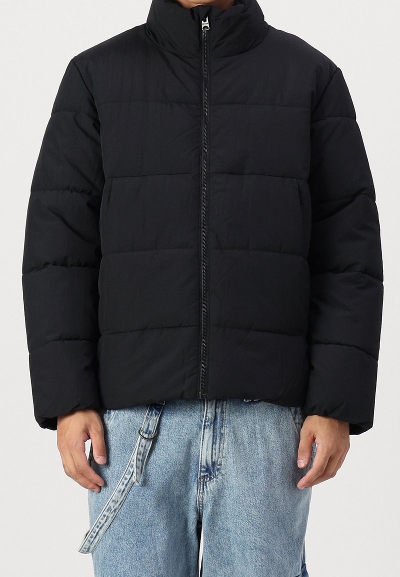 Chaqueta acolchada negra con cuello alto, cierre de cremallera y diseño acolchado. Presenta un ajuste holgado y está hecha de una tela suave e aislante.