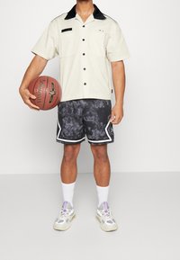 Camicia a maniche corte crema con colletto nero, abbinata a pantaloni corti neri con motivo e dettagli bianchi, mentre tiene un pallone da basket.