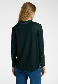 Tummanvihreä, pitkähihainen paita, jossa on sileä pinta. Varustettu kauluksella ja nappihihansuilla, takana hieman kaareva helma.