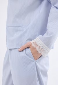 Conjunto de pijama a rayas azul claro con un bolsillo y bordado de encaje en los puños, que muestra una textura suave y un diseño de ajuste relajado.