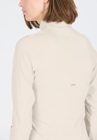 Helles beige Langarmshirt mit hohem Kragen, gerippter Textur und einem kleinen goldenen Akzent hinten in der Nähe der Schulternaht.