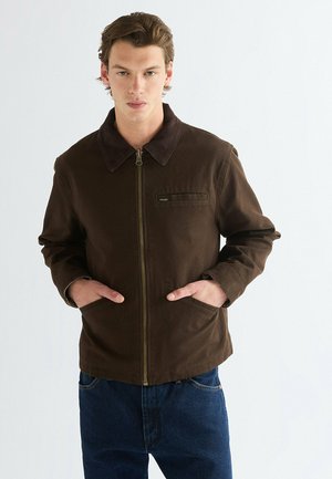 Junger Mann steht mit den Händen in den Taschen, trägt eine braune Zip-Jacke und blaue Jeans, schaut nach vorne vor einem schlichten, hellen Hintergrund.
