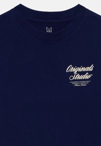 Tamsiai mėlyna medvilninė marškinėlių su apvalia iškirpte, priekyje su balta tekstine žyme "Originals Studio" ir mažą grafika krūtinės srityje.