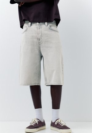 Mężczyzna ubrany w jasnoszare jeansowe szorty, ciemnofioletowe sneakersy z białymi podeszwami, białe skarpetki oraz ciemną koszulę z założonymi rękami.