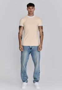 Beige overhemd met witte kraag en mouwen, denimblauwe jeans en witte sneakers; het model heeft tatoeages en staat tegen een grijze achtergrond.