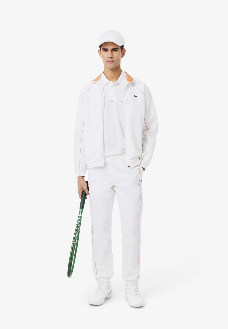 Junger Mann in weißer Sportkleidung und Cap, der einen grünen Tennisschläger hält und vor einem schlichten weißen Hintergrund steht.