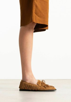 Jambe portant une mocassin en daim marron avec franges et pompons, associée à une jupe marron longueur genou, debout sur un sol gris clair.