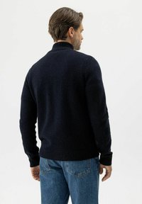 Mann mit braunem Haar, der einen schwarzen Strickpullover und blaue Jeans trägt, mit dem Rücken zu einem schlichten weißen Hintergrund.