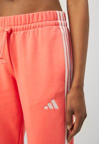 Koralne trenirke z belimi stranicami, elastičnim pasom, vrvico in Adidas logotipom na nogi. Mehka tkanina in sproščen kroj.