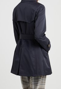 Žena v námořnickém trenčkotu s páskem a detailem na zadním yoku, spárovaném s hnědými a černými kostkovanými kalhotami, stojící před jednobarevným pozadím.