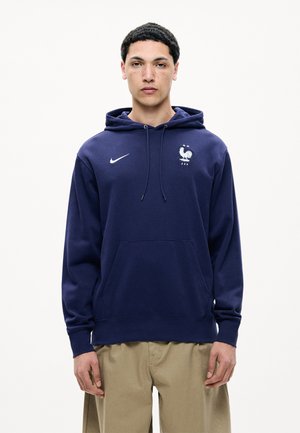 Ung mand med kort krøllet hår iført en mørkeblå hættetrøje med det franske fodboldforbunds logo og beige bukser, stående mod en hvid baggrund.