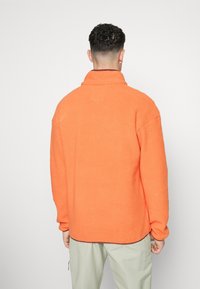 Hombre de espaldas que lleva una chaqueta de forro polar naranja y pantalones beige claros frente a un fondo blanco liso.