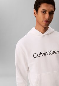Jauns vīrietis ar īsiem cirtainiem matiem, tērpies baltā kapucjakā ar "Calvin Klein" logotipu, stāvot pret pelēku fonu.