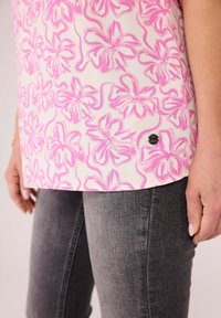 Ärmelloses Oberteil mit floralem Muster, das ein pinkes Blumenmuster auf crèmefarbenem Hintergrund zeigt, mit einem abgerundeten Saum und einem kleinen schwarzen Logoetikett an der Seite.