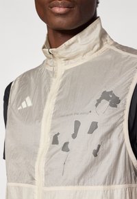 Ελαφρύ γιλέκο κρεμ χρώματος με ψηλό γιακά, υφή υφάσματος και μπροστινό φερμουάρ. Διαθέτει γκρι γραφικό σχέδιο.