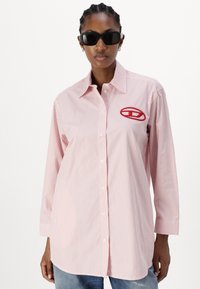 Striped long-sleeve shirt in light pink and white, featuring a button-down front and red embroidered logo on the left side.